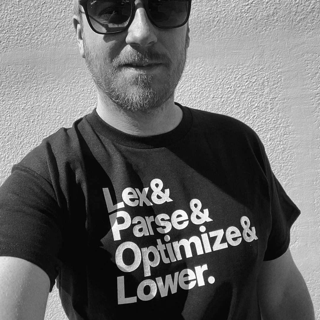 A photo of Rasmus in black and white looking great wearing shades and this t-shirt "Lex & Parse & Optimize & Lower."